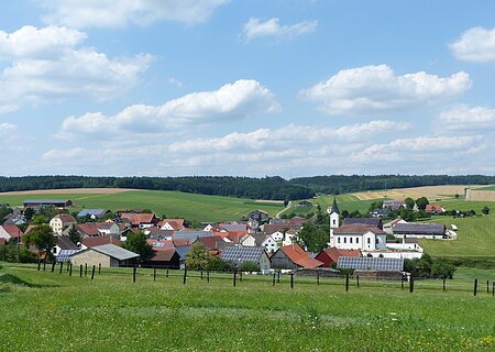 Aufnahme des kleines Dorfes Wittesheim mit roten Dächern und einer Kirche, umgeben von grünen Feldern und Wäldern unter blauem Himmel mit Wolken. Vorderhalb ein eingezäunter Bereich.