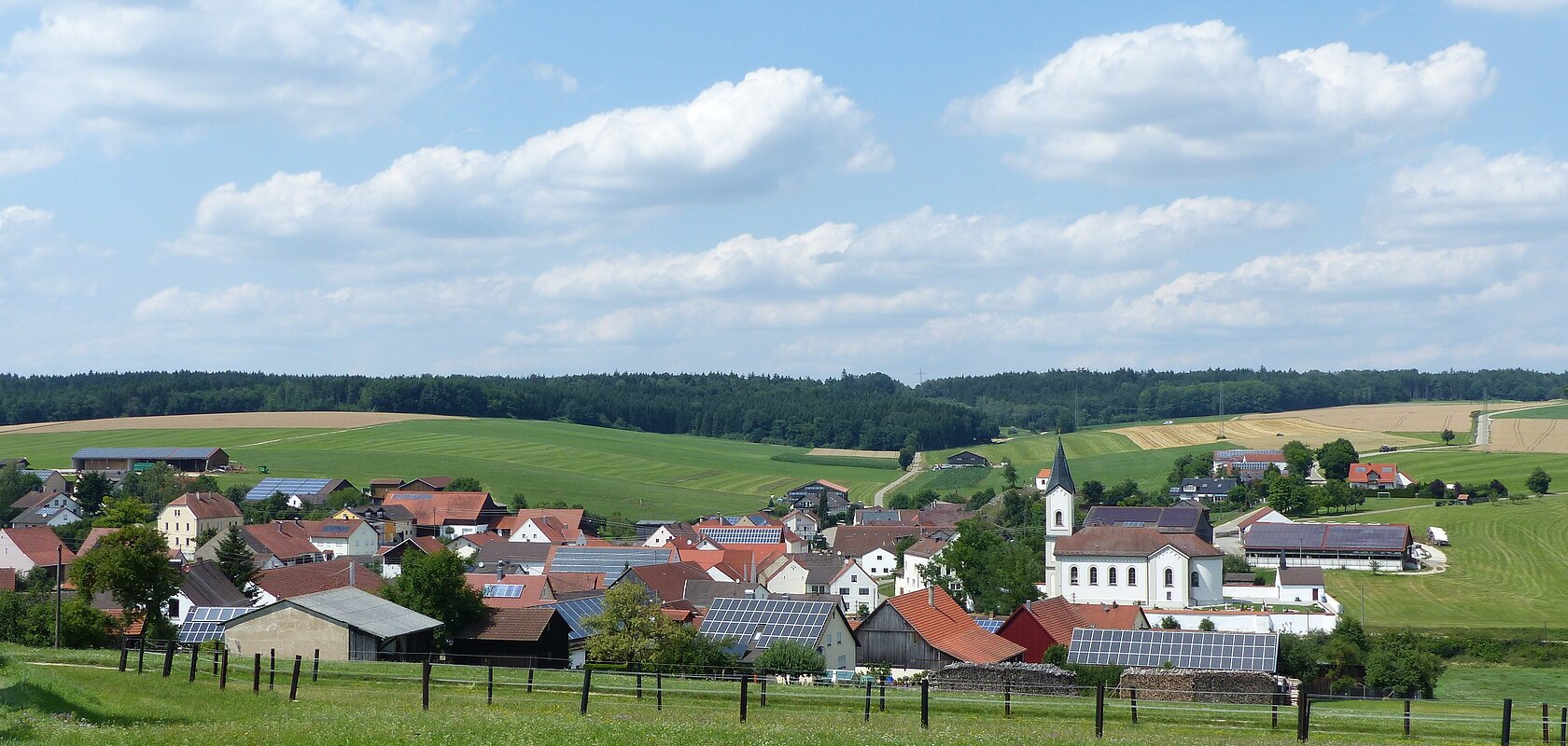 Aufnahme des kleines Dorfes Wittesheim mit roten Dächern und einer Kirche, umgeben von grünen Feldern und Wäldern unter blauem Himmel mit Wolken. Vorderhalb ein eingezäunter Bereich.