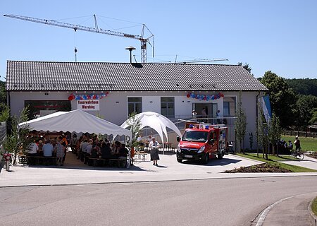 Feuerwehrhaus mit einem weißen Festzelt, einem weißen Pavillon und einem Feuerwehrfahrzeug davor. Menschen sitzen unter dem Zelt, ein Kran steht hinter dem Feuerwehrhaus, im Vordergrund eine gepflasterte Straße. Feuerwehrhaus ist umgeben von Feldern und einem Wald.