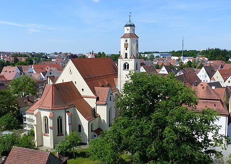 Luftbild der Stadtpfarrkirche mit Turm in einer Stadtlandschaft, umgeben von Häusern mit roten Dächern und Bäumen. Im Hintergrund blauer Himmel.