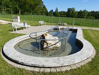 Kneipp-Gesundheitsanlage Monheim - Tretbecken Rundes Kneippbecken in Monheim mit einem Geländer und einem Barfußpfad, in der Mitte ein großer weißer Stein. Umgeben von einer Wiese, darauf Sitzmöglichkeiten und eine große Grünfläche mit Bäumen hinterhalb, abgetrennt mit einem Zaun.