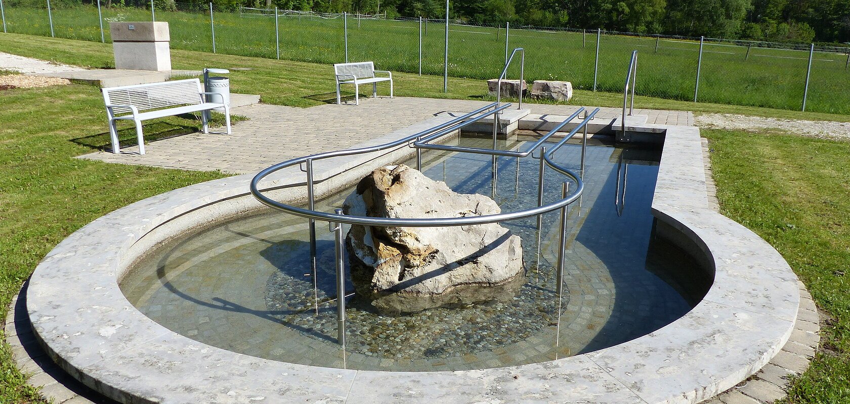 Rundes Kneippbecken in Monheim mit einem Geländer und einem Barfußpfad, in der Mitte ein großer weißer Stein. Umgeben von einer Wiese, darauf Sitzmöglichkeiten und eine große Grünfläche mit Bäumen hinterhalb, abgetrennt mit einem Zaun.
