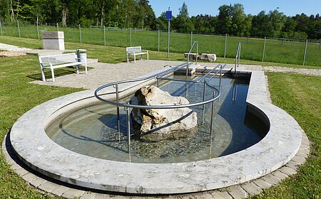 Rundes Kneippbecken in Monheim mit einem Geländer und einem Barfußpfad, in der Mitte ein großer weißer Stein. Umgeben von einer Wiese, darauf Sitzmöglichkeiten und eine große Grünfläche mit Bäumen hinterhalb, abgetrennt mit einem Zaun.