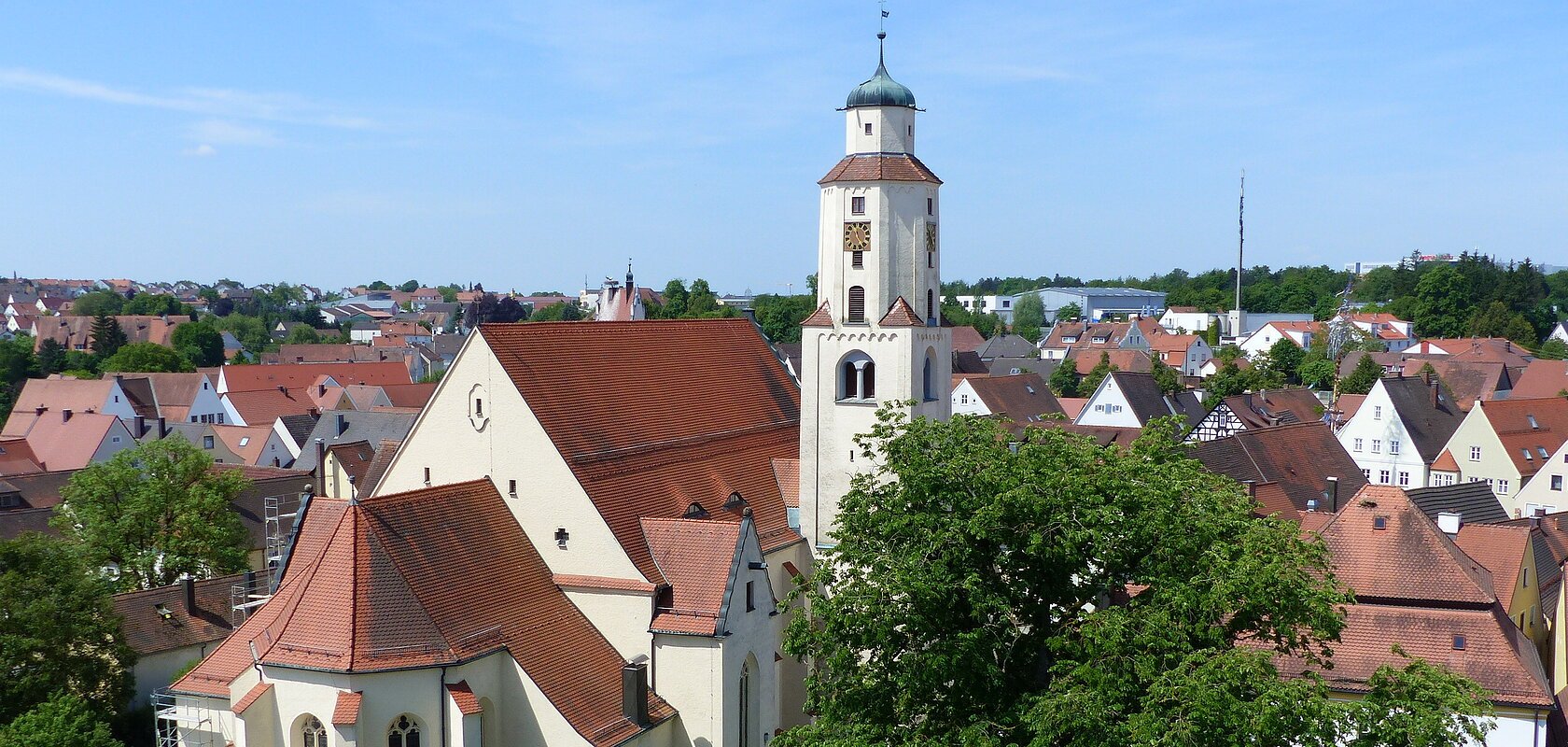 Luftbild der Stadtpfarrkirche mit Turm in einer Stadtlandschaft, umgeben von Häusern mit roten Dächern und Bäumen. Im Hintergrund blauer Himmel.