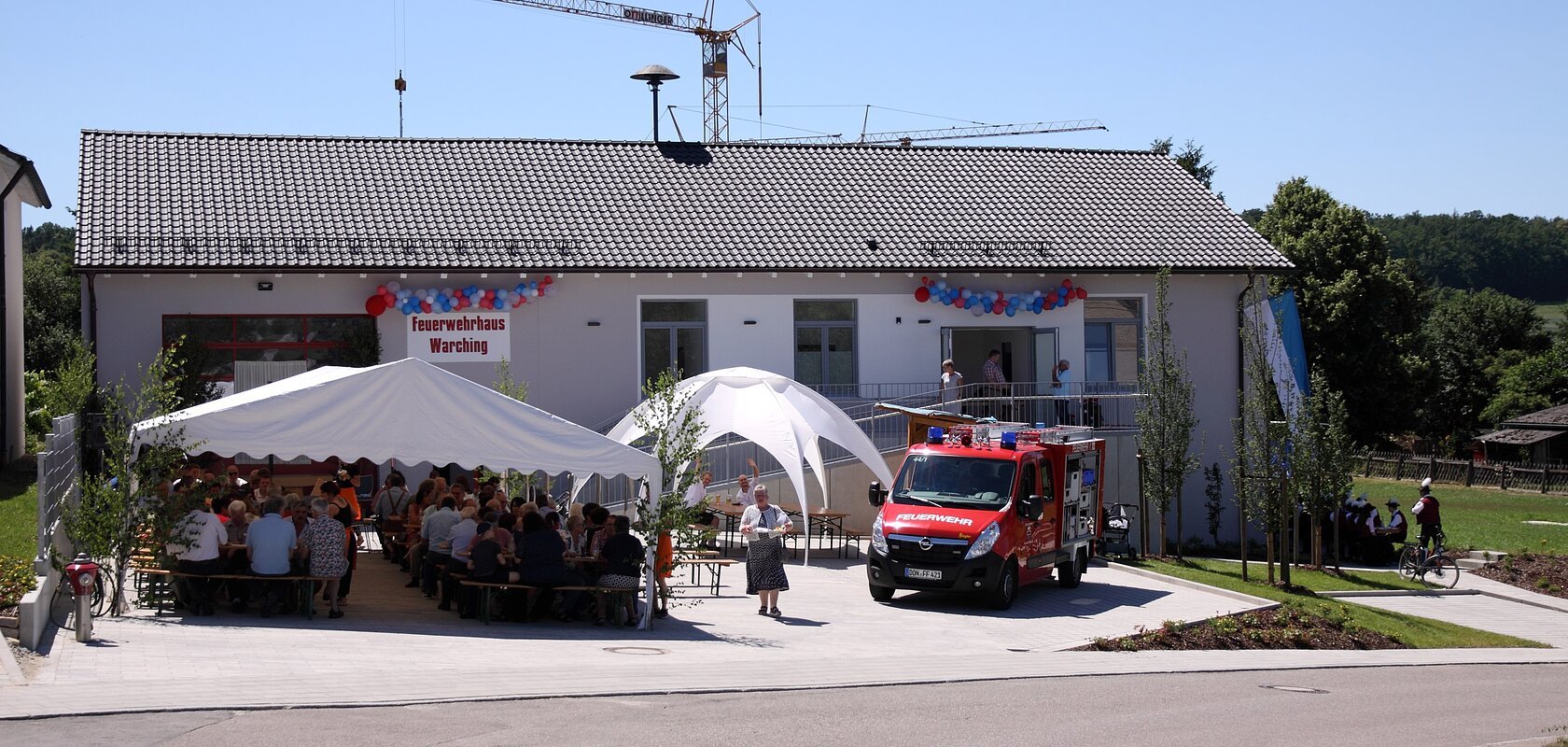 Feuerwehrhaus mit einem weißen Festzelt, einem weißen Pavillon und einem Feuerwehrfahrzeug davor. Menschen sitzen unter dem Zelt, ein Kran steht hinter dem Feuerwehrhaus, im Vordergrund eine gepflasterte Straße. Feuerwehrhaus ist umgeben von Feldern und einem Wald.