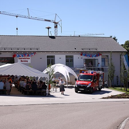 Feuerwehrhaus mit einem weißen Festzelt, einem weißen Pavillon und einem Feuerwehrfahrzeug davor. Menschen sitzen unter dem Zelt, ein Kran steht hinter dem Feuerwehrhaus, im Vordergrund eine gepflasterte Straße. Feuerwehrhaus ist umgeben von Feldern und einem Wald.