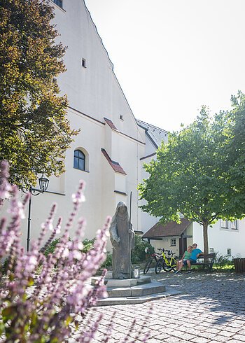 Die Walburgastatue in Monheim - im Stadtzentrum Steinfigur der heiligen Walburga auf einem gepflasterten Platz vor der Stadtpfarrkirche St. Walburga in Monheim, daneben zwei sitzende Personen auf einer Bank, dahinter ein Baum. Im Vordergrund unscharf Blumen.