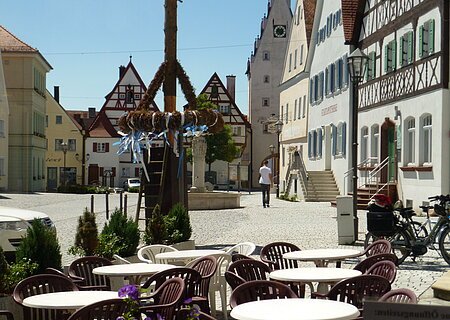 Ein Marktplatz mit Tischen und Stühlen eines Cafés im Vordergrund, Fachwerkhäusern im Hintergrund und einem Maibaum in der Mitte des Marktplatzes. Eine Person läuft auf dem Markplatz.