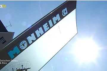Ein Schild mit der Aufschrift "Monheim" vor einem blauen Himmel und Sonnenschein. Links oben das Logo von a.tv. Im Hintergrund ein Haus.
