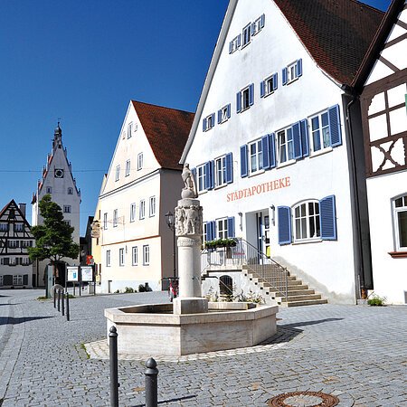 Historischer gepflasterter Marktplatz mit Fachwerkhäusern auf einem Gebäude steht die Aufschrift "Stadtapotheke". Ein Brunnen mit einer Steinskulptur steht davor. Links verläuft eine Straße.