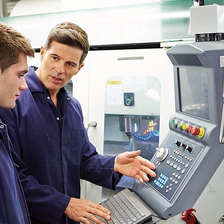 Zwei Personen in blauen Kitteln stehen vor einer CNC-Maschine und diskutieren die Bedienung.