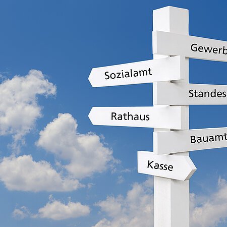 Weißer Wegweiser mit Schildern zu Sozialamt, Rathaus, Kasse, Gewerbeamt, Standesamt, Bauamt vor blauem Himmel mit Wolken.