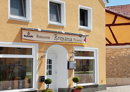 Gelbes Gebäude mit mehreren Fenstern und einem Schild "Ristorante Romana Pizzeria" über dem Eingang. Zwei kleine Bäume links und rechts vor der Tür.