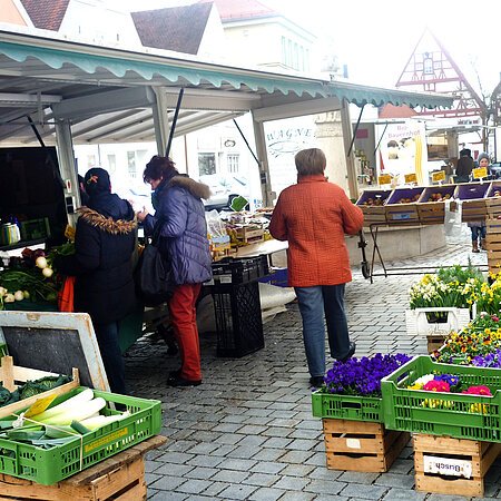 Menschen kaufen an einem Marktstand mit Gemüse und Blumen auf einem gepflasterten Platz ein.
