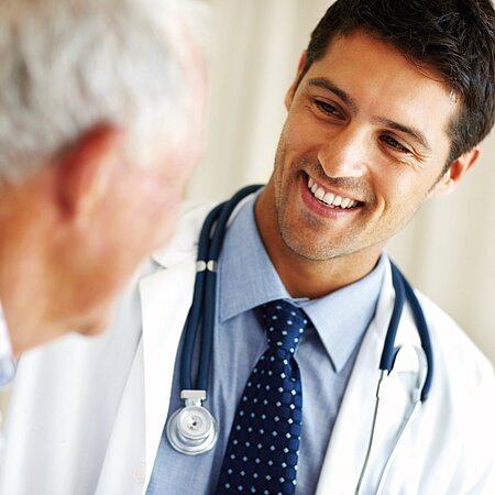 Ein Arzt im weißen Kittel mit einem Stethoskop um den Hals spricht lächelnd mit einem älteren Mann.