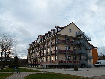 Donau-Ries Seniorenheim Monheim Mehrstöckiges orange gestrichenes Gebäude mit vielen Fenstern, Balkonen und einer großen Außentreppe, umgeben von Gehwegen und Rasenfläche. Vorderhalb ein gepflasterter Platz mit einem großen Stein drauf. Links im Bild eine Sitzbank. Im Hintergrund bewölkter Himmel.