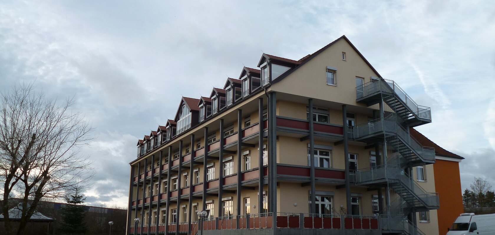 Mehrstöckiges orange gestrichenes Gebäude mit vielen Fenstern, Balkonen und einer großen Außentreppe, umgeben von Gehwegen und Rasenfläche. Vorderhalb ein gepflasterter Platz mit einem großen Stein drauf. Links im Bild eine Sitzbank. Im Hintergrund bewölkter Himmel.