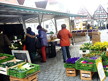 Wochenmarkt Monheim Menschen kaufen an einem Marktstand mit Gemüse und Blumen auf einem gepflasterten Platz ein.