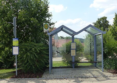 Bushaltestelle mit Glasüberdachung, umgeben von Bäumen und Sträuchern, mit Fahrplan und Haltestellenschild.
