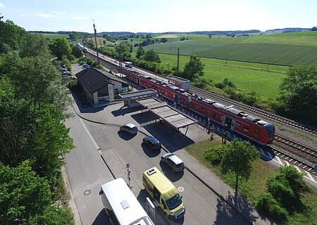 Luftaufnahme eines Bahnhofs mit rotem Zug, umgeben von Feldern und Bäumen. Stehende Autos und Busse sind zu sehen.
