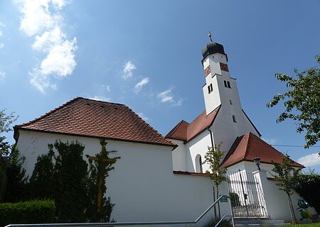 Weiße Kirche mit rotem Ziegeldach und Zwiebelturm vor blauem Himmel. Im Vordergrund ein Tor und Pflanzen.