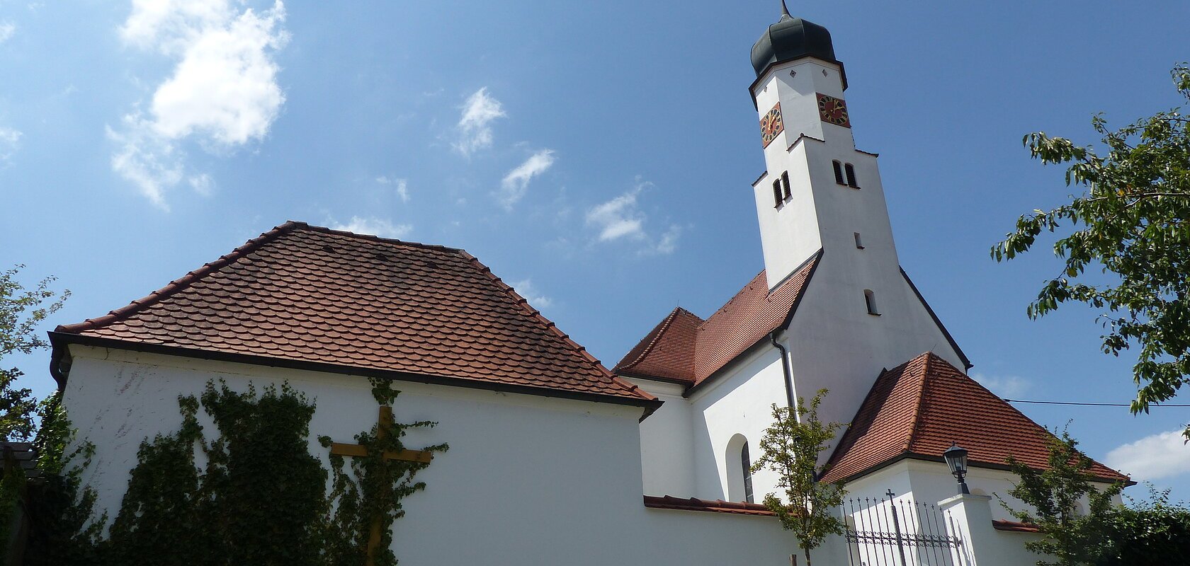 Weiße Kirche mit rotem Ziegeldach und Zwiebelturm vor blauem Himmel. Im Vordergrund ein Tor und Pflanzen.