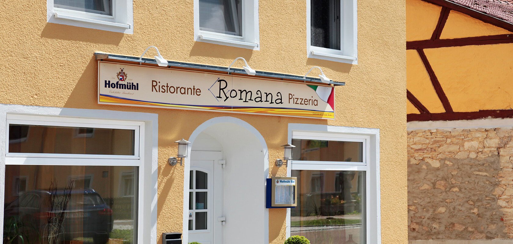 Pizzeria Ristorante Romana - Mediterraner Charme in Monheim Gelbes Gebäude mit mehreren Fenstern und einem Schild "Ristorante Romana Pizzeria" über dem Eingang. Zwei kleine Bäume links und rechts vor der Tür.
