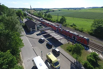 Luftaufnahme eines Bahnhofs mit rotem Zug, umgeben von Feldern und Bäumen. Stehende Autos und Busse sind zu sehen.
