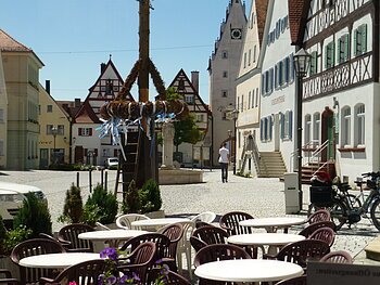 Café Konditorei Wenninger - das Café am Marktplatz Ein Marktplatz mit Tischen und Stühlen eines Cafés im Vordergrund, Fachwerkhäusern im Hintergrund und einem Maibaum in der Mitte des Marktplatzes. Eine Person läuft auf dem Markplatz.
