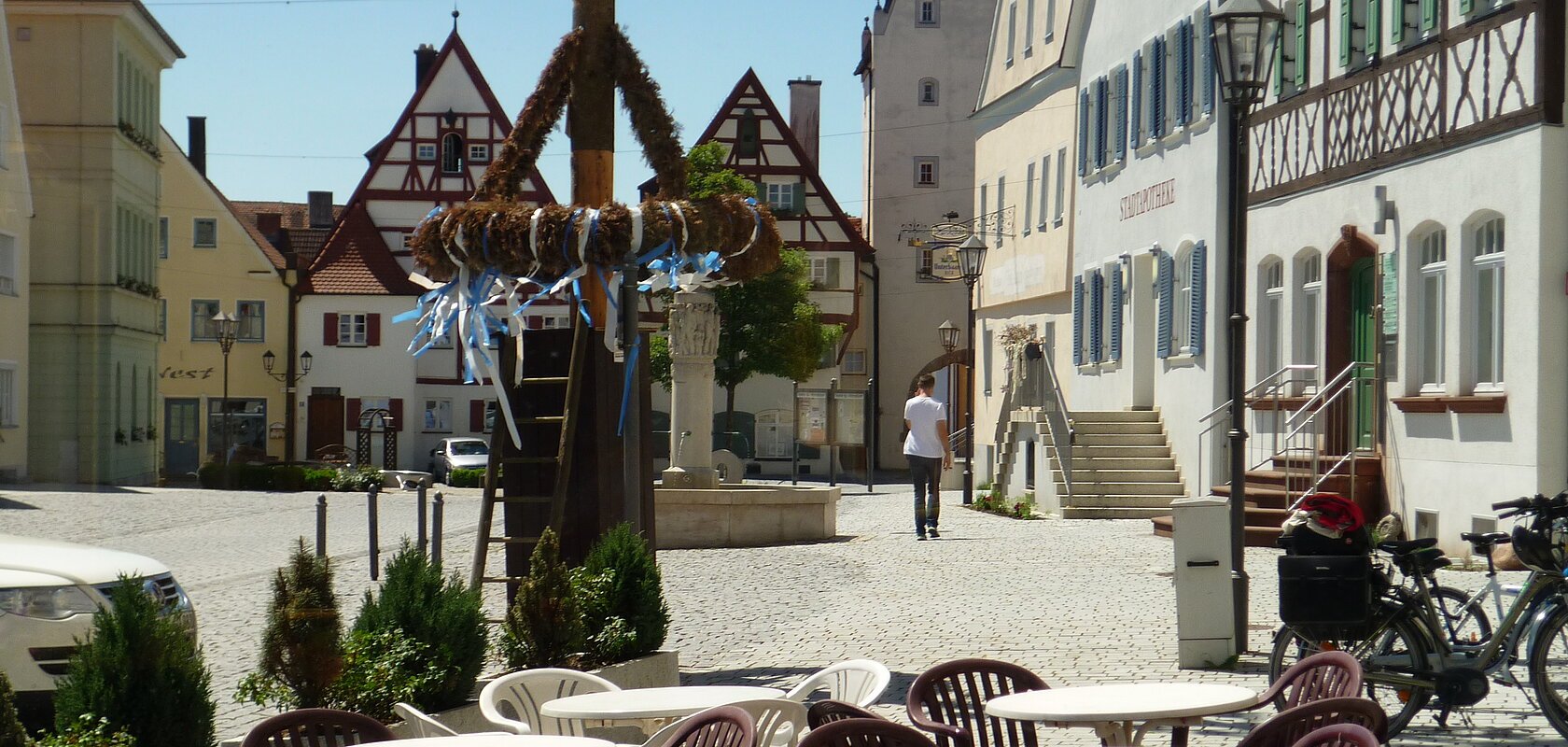 Ein Marktplatz mit Tischen und Stühlen eines Cafés im Vordergrund, Fachwerkhäusern im Hintergrund und einem Maibaum in der Mitte des Marktplatzes. Eine Person läuft auf dem Markplatz.
