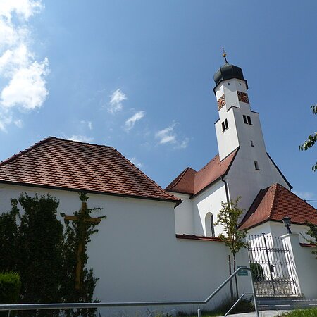 Weiße Kirche mit rotem Ziegeldach und Zwiebelturm vor blauem Himmel. Im Vordergrund ein Tor und Pflanzen.