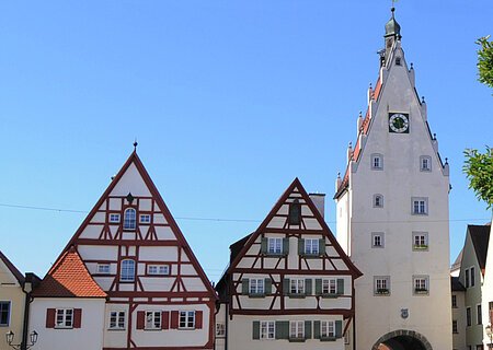Historische Fachwerkhäuser, ein Torturm mit Uhr in einer Altstadt bei klarem Himmel. Links im Bild parkende Autos auf einem gepflasterten Platz. Rechts im Vordergrund ein Baum mit einer Info-Tafel und einem großen Buchstaben "O" aus Stein. Dahinter eine Informationsvetrine.