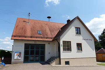Ein Feuerwehrgebäude mit der Aufschrift "Feuerwehr Itzing" mit einem roten Ziegeldach, zwei Fenstern, einer Treppe und einem großen Tor. Im Hintergrund blauer Himmel und Bäume. Auf dem Gebäude ist ein Zigarettenautomat angebracht.