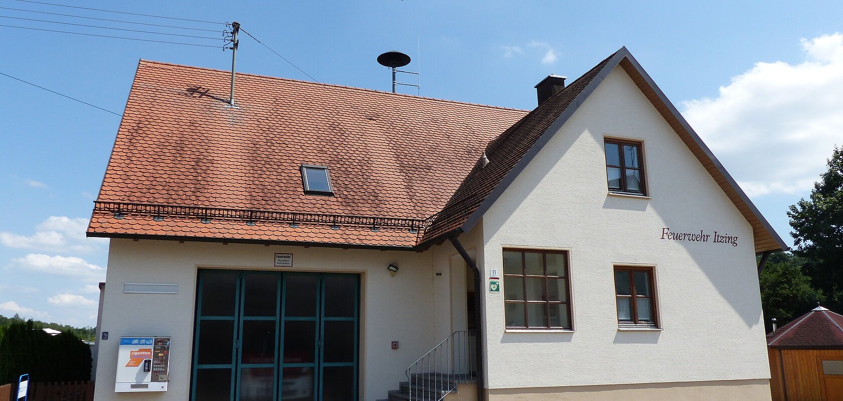 Ein Feuerwehrgebäude mit der Aufschrift "Feuerwehr Itzing" mit einem roten Ziegeldach, zwei Fenstern, einer Treppe und einem großen Tor. Im Hintergrund blauer Himmel und Bäume. Auf dem Gebäude ist ein Zigarettenautomat angebracht.
