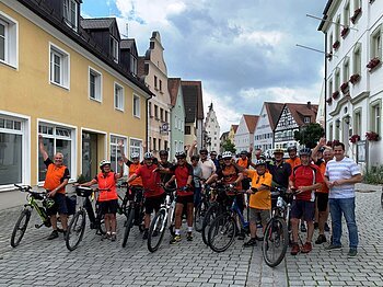 "Anradeln" - Stadtradeln Monheim 2024 Gruppe von Radfahrern in bunter Sportkleidung posiert mit Fahrrädern auf einer gepflasterten Straße in einer Stadt.
