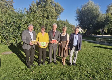 Fünf Personen stehen auf einer Wiese vor Bäumen, drei halten bunte Geschenke in den Händen, im Hintergrund blauer Himmel. Renteneintritt der drei langjährigen Mitarbeiter der VG Monheim.
