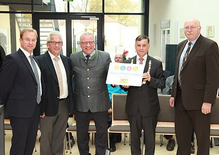 Fünf Männer in Anzügen stehen in einem Raum, einer hält ein Schild mit der Aufschrift "Bayern barrierefrei". Im Hintergrund große Eingangstür aus Glas.