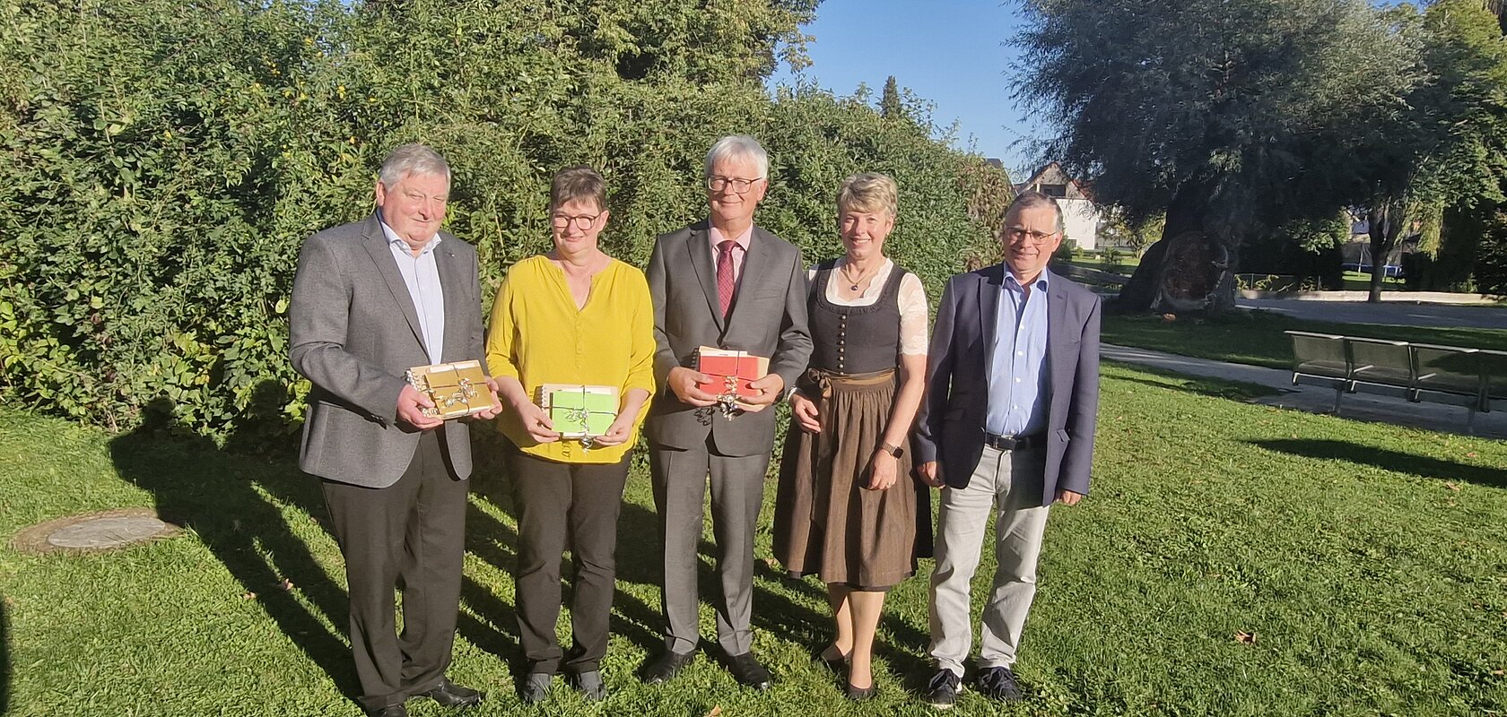 Fünf Personen stehen auf einer Wiese vor Bäumen, drei halten bunte Geschenke in den Händen, im Hintergrund blauer Himmel. Renteneintritt der drei langjährigen Mitarbeiter der VG Monheim.