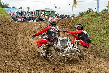 Zwei Personen fahren ein Motocross-Gespann auf einer schlammigen Strecke, im Hintergrund sind Zuschauer.