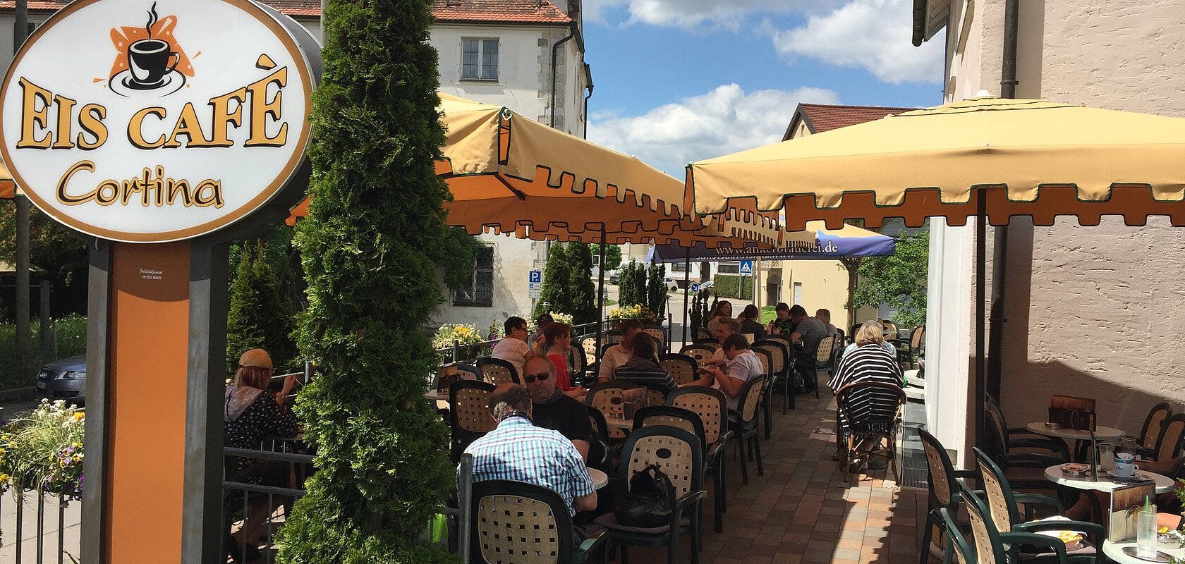 Außenbereich eines Eiscafés mit gelben Sonnenschirmen, Tischen und Stühlen, Gäste sitzen und essen.