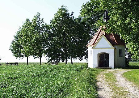 Kleine Kapelle mit rotem Dach auf einer Wiese mit einem Schotterweg. Umgeben von einem grünem Feld und Bäumen.