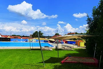 Ein Freibad mit Schwimmbecken, Sonnensegeln, Sitzmöglichkeiten und einem Sandkasten auf einer grünen Wiese, rechts unten im Bild. Im Hintergrund blauer Himmel mit Wolken.