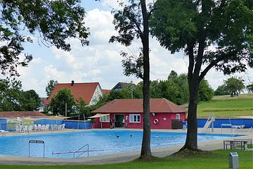 Ein Freibad mit Schwimmbecken, einer Rutsche und Sitzmöglichkeiten, umgeben von Bäumen und Wiesen. Im Hintergrund steht ein rotes Gebäude mit Fenstern, ein Kiosk.