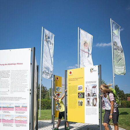 Eine erwachsene Personen und ein Kind stehen auf einer gepflasterten Fläche neben Informationstafeln im Freien, umgeben von Fahnen. Im Hintergrund grüne Wiese und blauer Himmel.