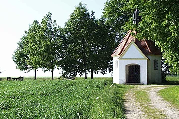 Kleine Kapelle mit rotem Dach auf einer Wiese mit einem Schotterweg. Umgeben von einem grünem Feld und Bäumen.