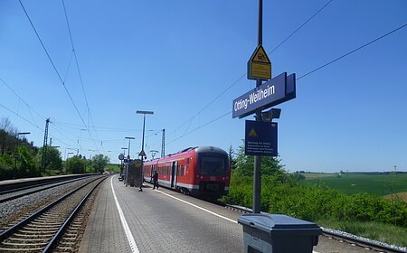 Der Bahnhof Otting-Weilheim mit rotem Zug auf dem Gleis, blauer Himmel, grüne Landschaft im Hintergrund.