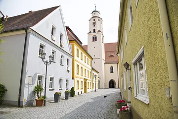 Stadtpfarrkirche Monheim Ansicht einer alten Innenstadt mit bunten Häusern und einem Kirchturm im Hintergrund. Vor einigen Häusern stehen Pflanzgefäße und eine kleine Katze läuft auf der Straße.