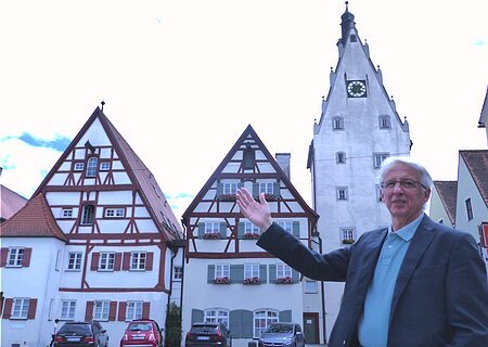Eine Person mit Brille, im Sakko und blauem Hemd steht vor Fachwerkhäusern und einem Turm mit Uhr und zeigt darauf. Vor den Häusern stehen auf einer gepflasterten Fläche vier Autos geparkt.