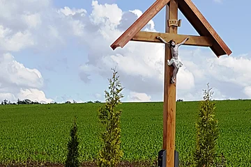 Flurkreuz am Wegesrand zwischen Monheim und Warching Holzkreuz mit überdachter Christusfigur mit bepflanztem Beet darunter, daneben eine Holzbank. Im Hintergrund ein Feld und blauer Himmel.