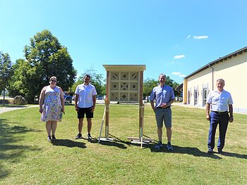 Wildbienen Nisthilfe Vier Personen stehen auf einer Wiese links und rechts neben einem großen Insektenhotel, im Hintergrund Bäume, blauer Himmel und ein weiß gestrichenes Gebäude.